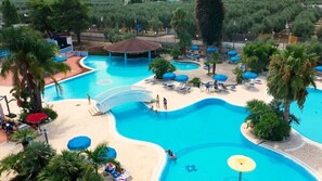A large outdoor swimming pool with a winding design, surrounded by palm trees and sun loungers. - Villaggio Merino (Vieste)
