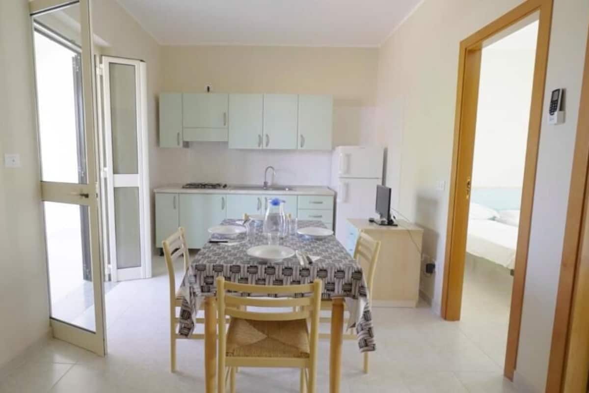 A small, bright kitchen with light green cabinets, a dining table set with plates and glasses, and a view of the sea through an open door.