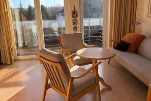 Ein Zimmer mit einer Couch, zwei Sesseln, einem kleinen Tisch und Blick durch eine Glastür nach draußen.