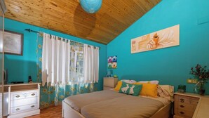 Een slaapkamer met een houten plafond, een bed met veel kussens, een nachtkastje, een dressoir en een schilderij aan de muur.