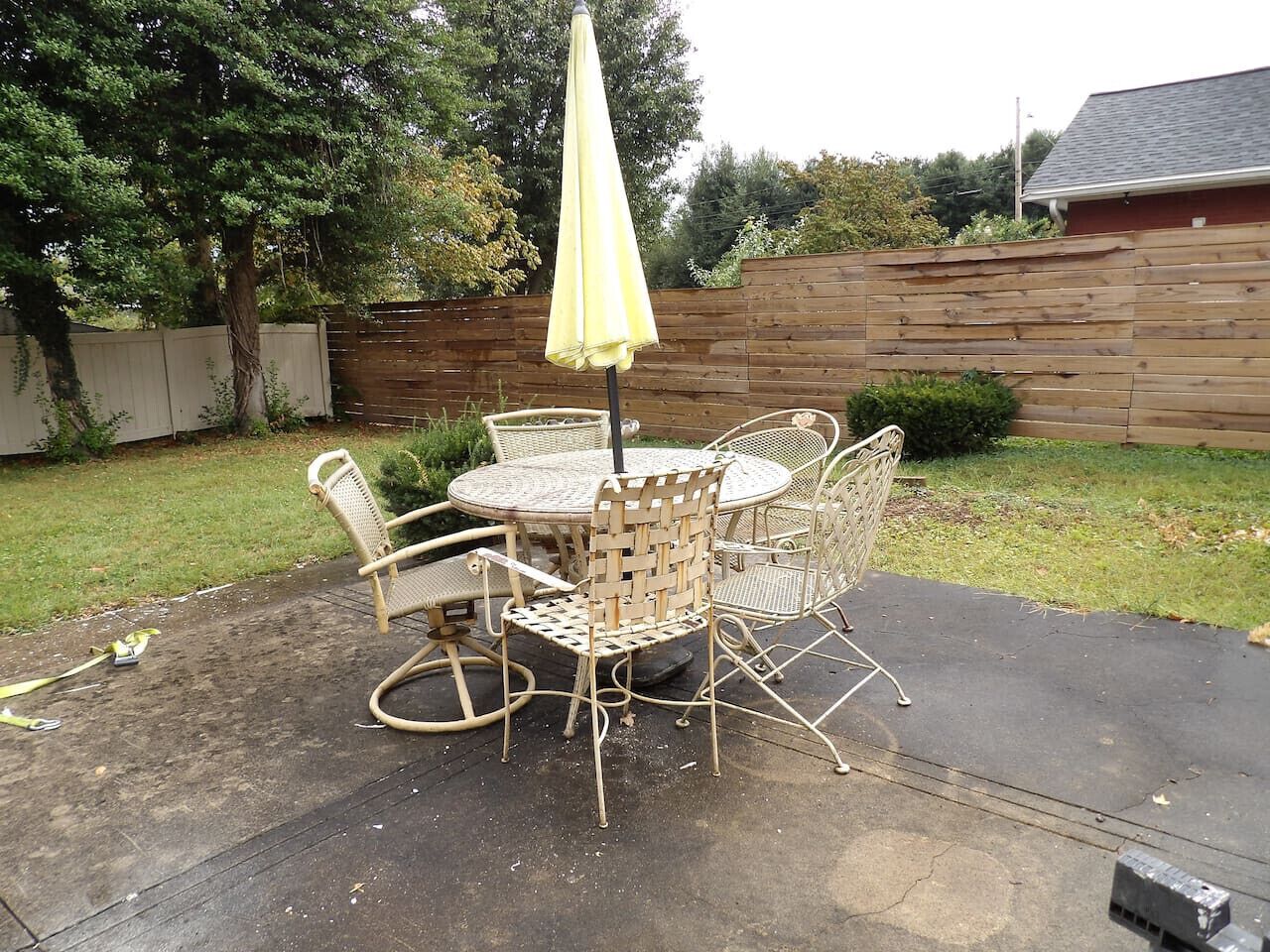 An outdoor patio with a round table and four wicker chairs, an umbrella, and a wooden fence in the background.