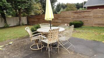 An outdoor patio with a round table and four wicker chairs, an umbrella, and a wooden fence in the background.