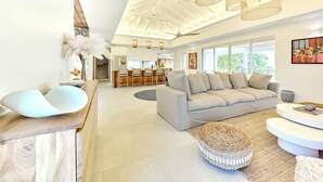 Ein geräumiger Wohnbereich mit einem modernen Sofa, einem Holztisch und einem gewebten Hocker. Im Hintergrund ist ein Essbereich mit Holzstühlen und einem großen Fenster zu sehen.