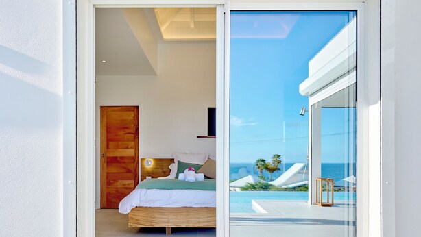 Ein Schlafzimmer mit Holztür, einem Bett mit weißer Bettwäsche und Blick auf ein Schwimmbad und Meer durch eine Glastür.