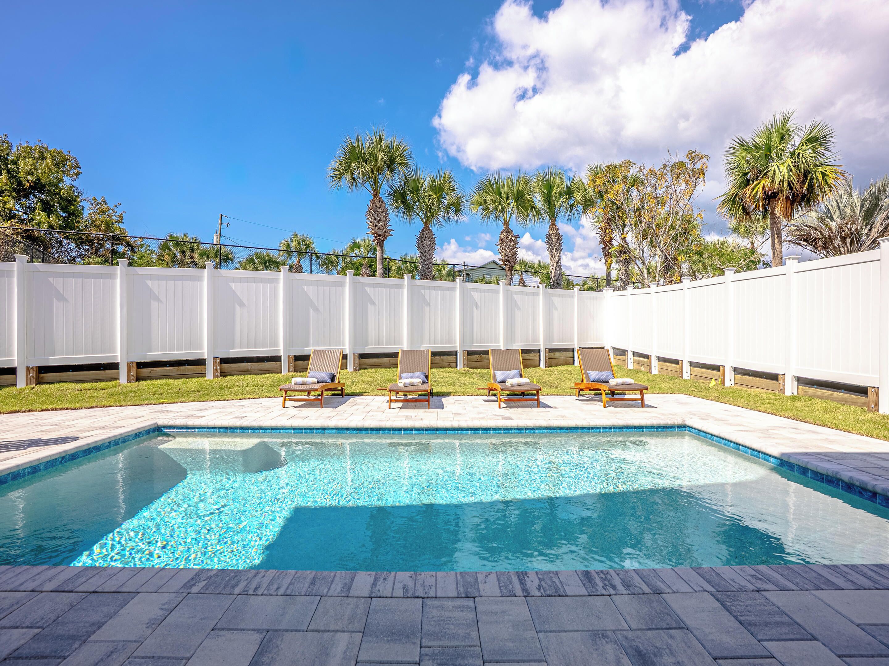 A swimming pool with four lounge chairs, a white fence, and palm trees in the background.
