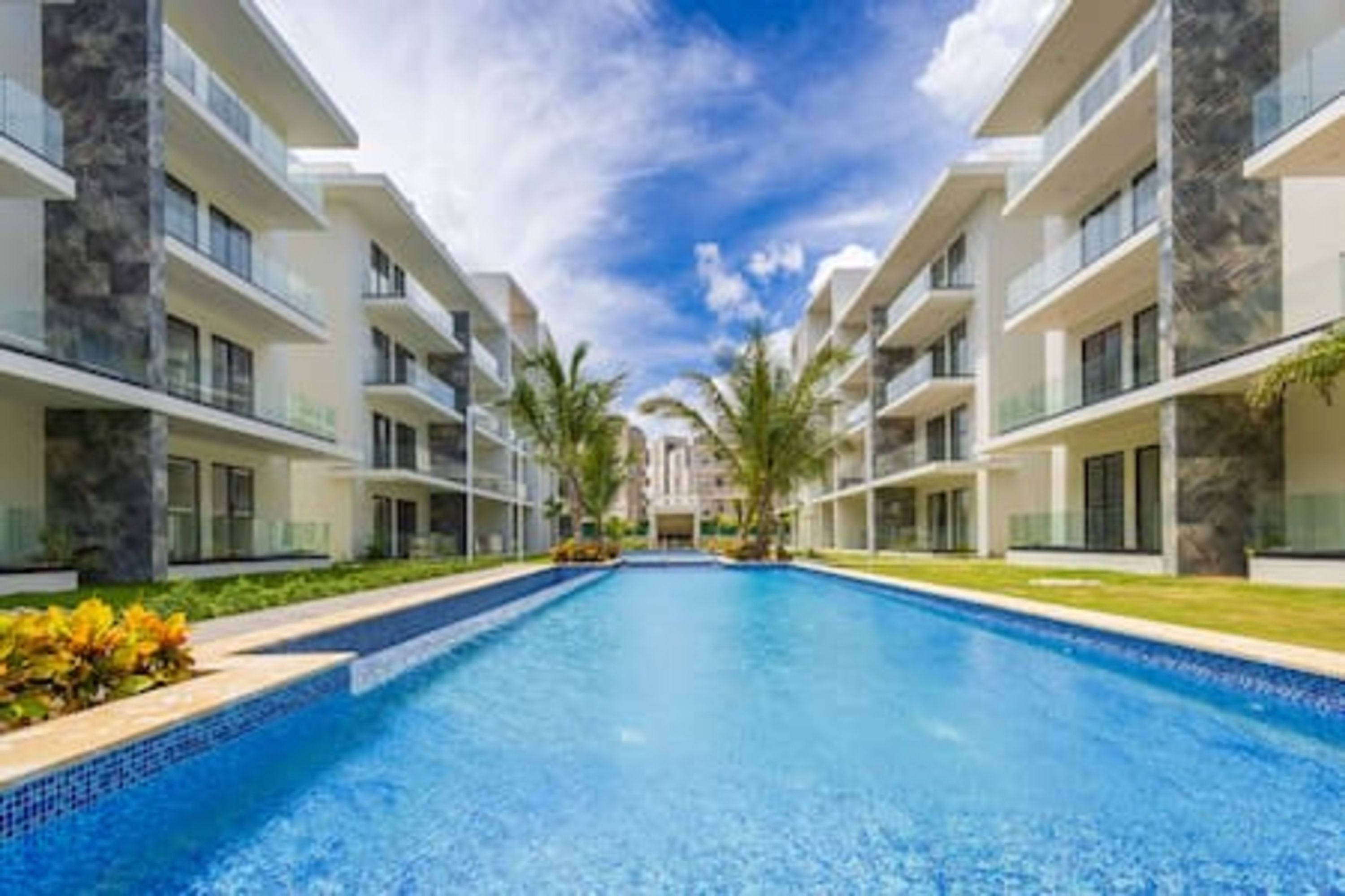 A modern hotel with a central swimming pool, multiple balconies, and a clear blue sky.