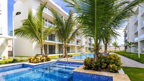 Une piscine aux bords bleus recouverts de carreaux, entourée de palmiers et bordée d’un bâtiment blanc doté de balcons.