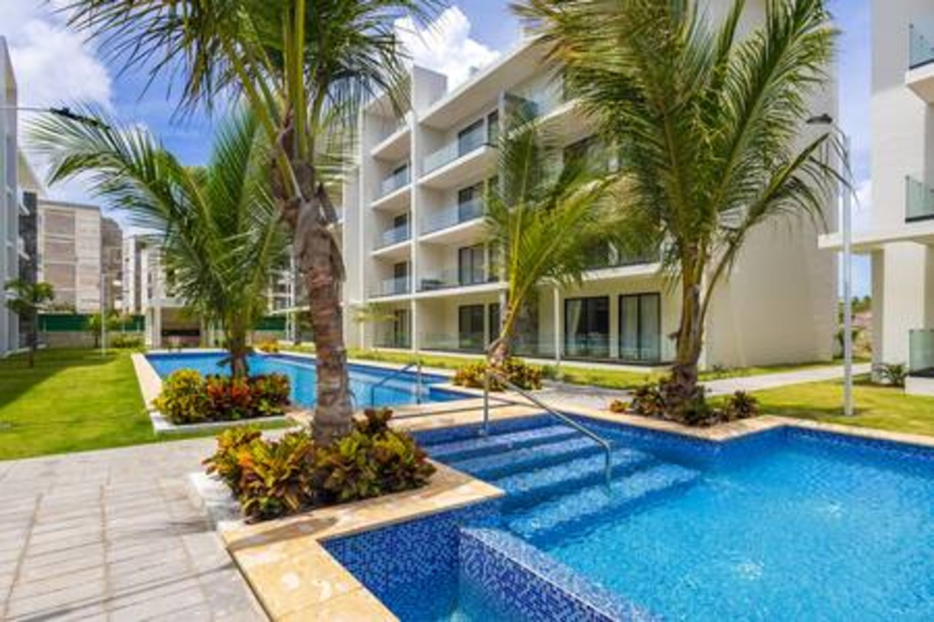 A modern hotel with a pool area, palm trees, and a clear blue sky.