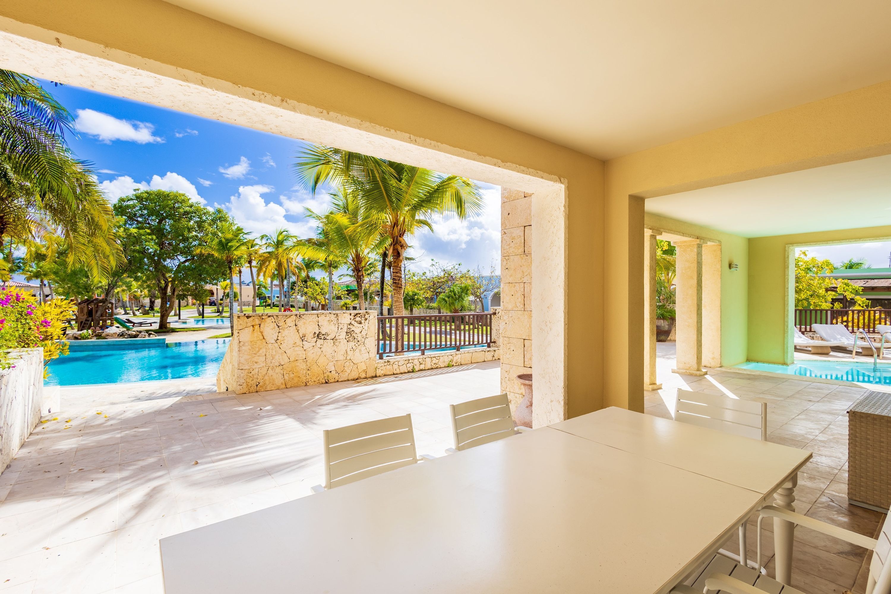 Zona junto a la piscina con una mesa y sillas blancas, pared de piedra y vistas a una pileta y palmeras.