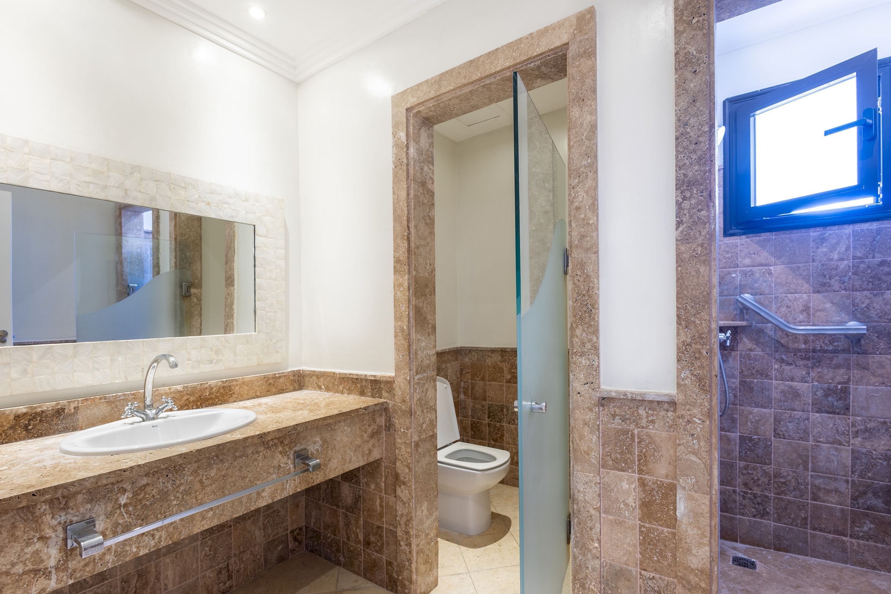 Un baño moderno con lavamanos de mármol, cabina de ducha con paredes de vidrio y un inodoro visible a través de una puerta de cristal.