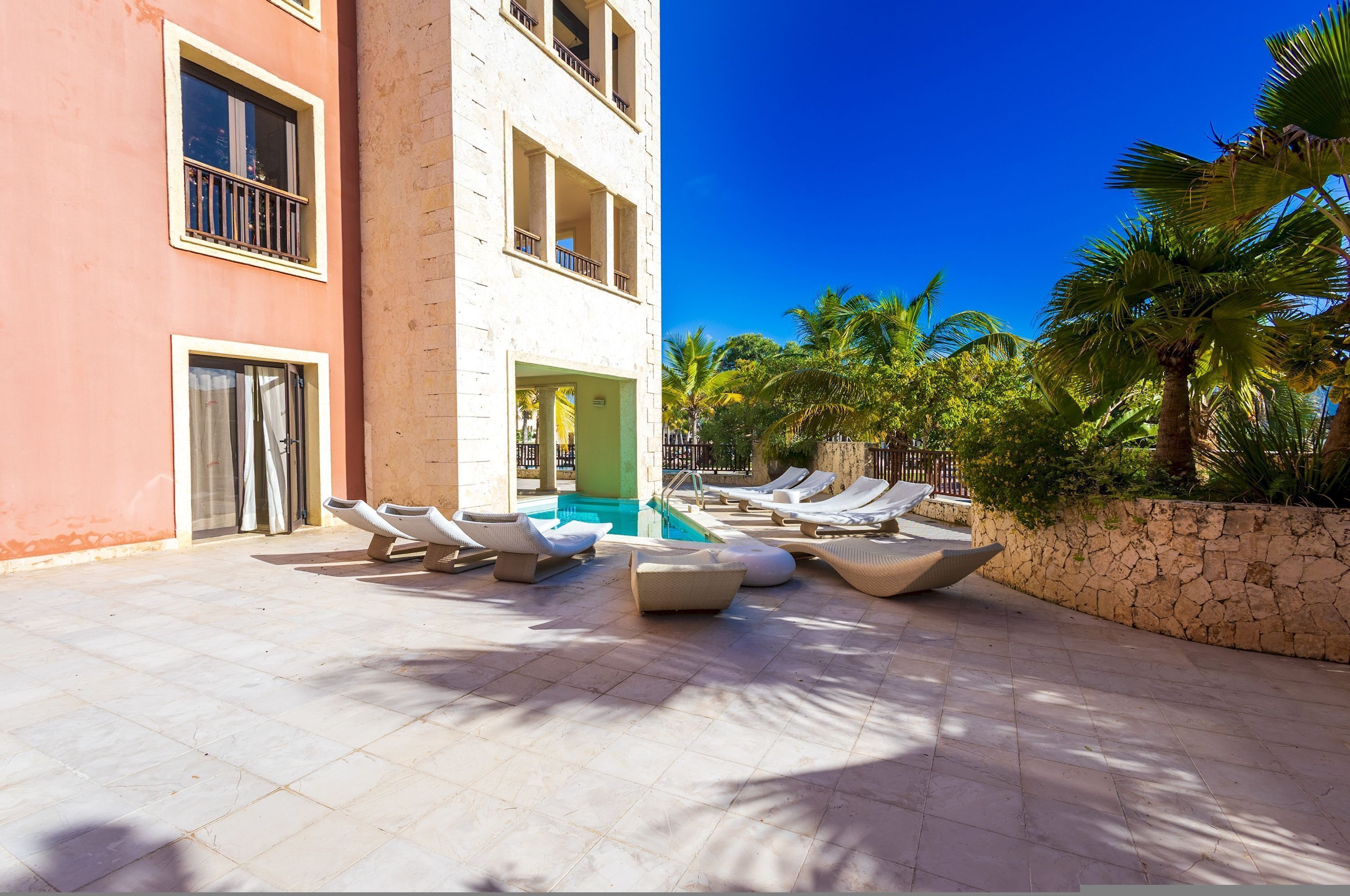 A poolside area with lounge chairs and a pool, surrounded by palm trees and a building with balconies.
