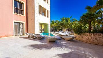A poolside area with lounge chairs and a pool, surrounded by palm trees and a building with balconies.