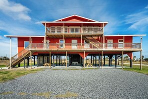 A two-story building with a balcony on the upper level, wooden railings, and a gravel driveway. - Big 6BR Beach Getaway for 20 | Near Ferry, 3 Units (Bolivar Peninsula)