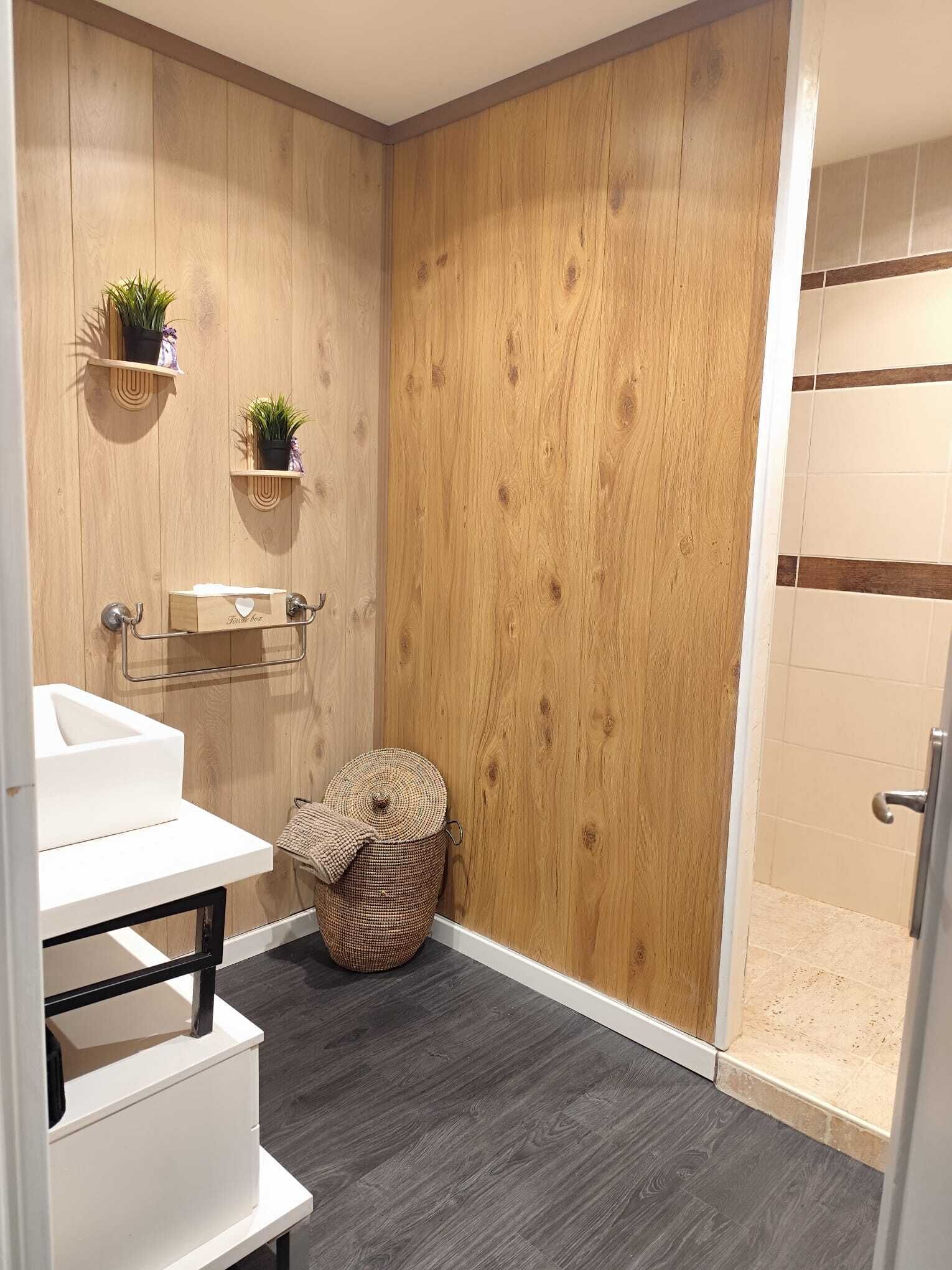 A bathroom with a wooden wall, a white sink, two shelves with plants, a round woven basket, and a shower area.
