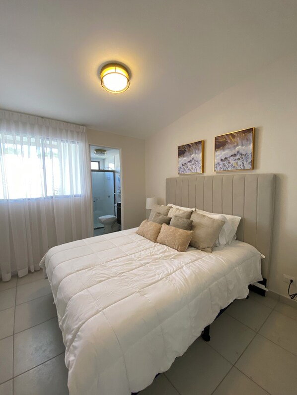 A bedroom with a large bed, a headboard, two wall-mounted paintings, and a ceiling light.