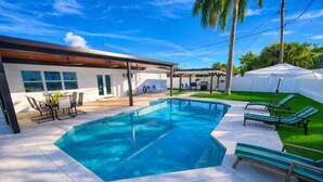 Une piscine rectangulaire entourée d’une terrasse carrelée, de chaises longues et d’un patio couvert.
