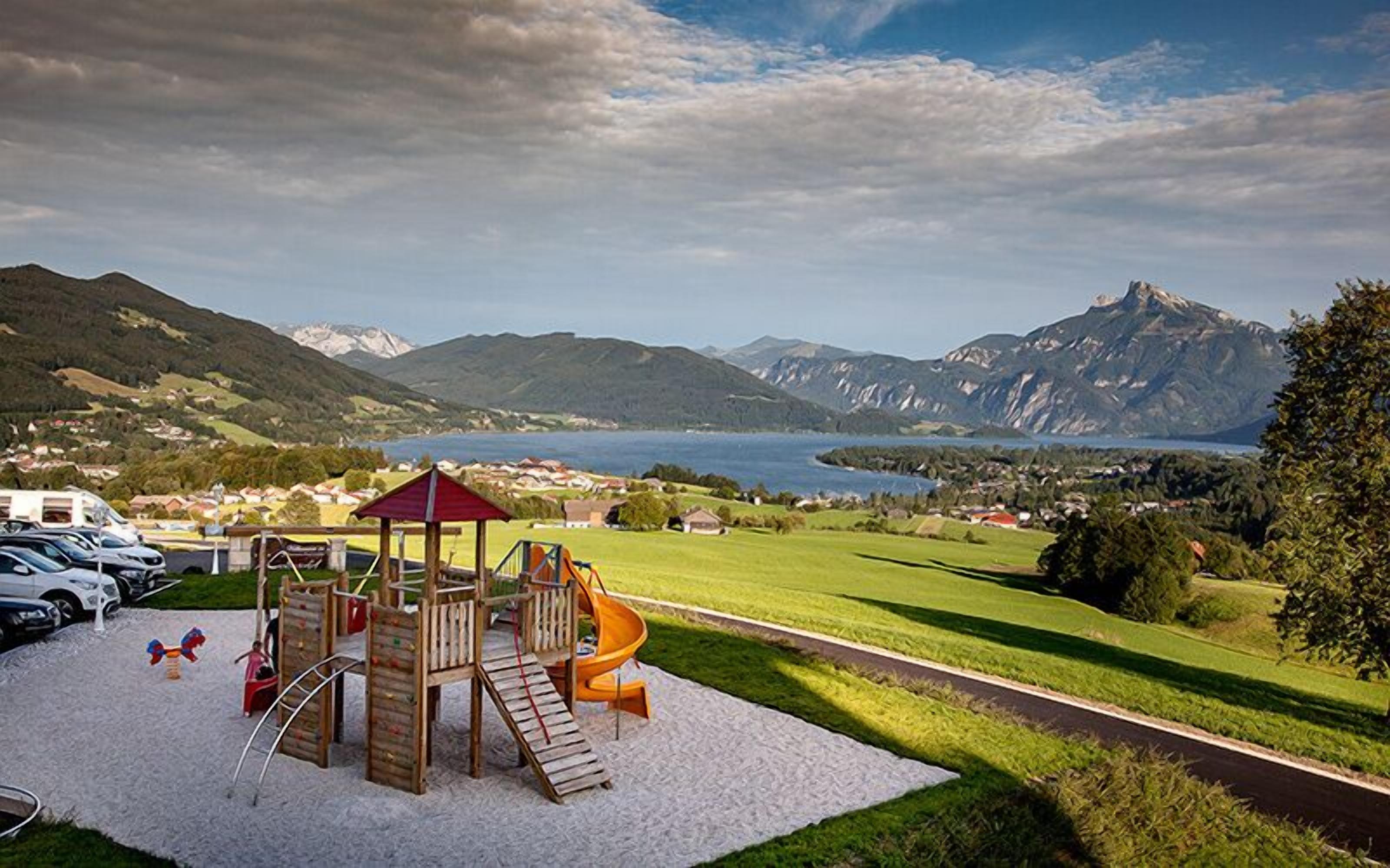 A playground with a wooden play structure and a slide, set against a backdrop of mountains and a lake.