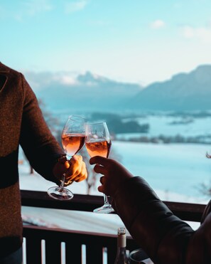 Deux personnes trinquent avec des verres à vin, une bouteille de vin est posée à proximité, et en arrière-plan, on aperçoit un paysage magnifique avec des montagnes et de l’eau.