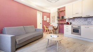Ein kompakter Wohnraum mit grauem Sofa, einem kleinen runden Tisch, einer Küchenzeile mit weißen Schränken und einer Mikrowelle sowie einem Esstisch mit Stühlen.
