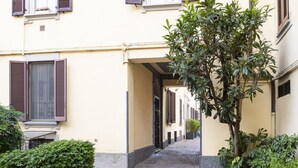 Ein schmaler Kopfsteinpflastergang zwischen Gebäuden mit Fensterläden, einem Baum und einem kleinen Busch.