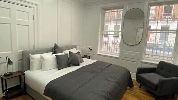 Un dormitorio con una cama grande, una silla, una mesita de noche y un ventanal con vistas a edificios.