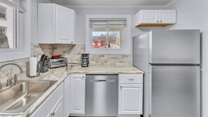 A modern kitchen with white cabinets, a stainless steel dishwasher, a refrigerator, a window with blinds, and a view of a red building outside. - Main Lineup Apt 4 - All Star Baseball Rentals (Oneonta)