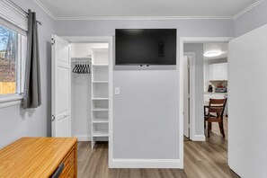 A room with a built-in TV, a wooden dresser, a closet with hanging space, and a kitchen area visible through an open door. - Main Lineup Apt 4 - All Star Baseball Rentals (Oneonta)