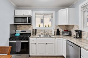 A compact kitchen with white cabinetry, a microwave, a stove, a sink, and a window with a view of buildings. - Main Lineup Apt 4 - All Star Baseball Rentals (Oneonta)