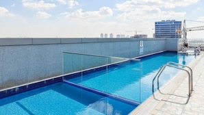A rooftop swimming pool with clear blue water, a glass barrier, and a stone deck. - Modern Studio with Balcony in Dubai Sports City (Dubai)