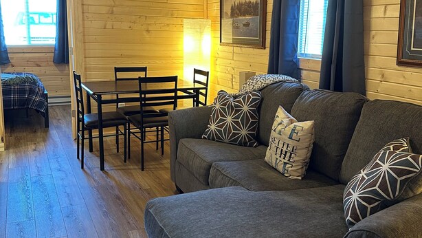 A cozy cabin interior with a gray sectional sofa, a dining table with chairs, wooden floors, a ceiling fan, and a framed picture on the wall. - Perfect 3-bedroom cabin with AC in pleasant Moravia (Moravia)