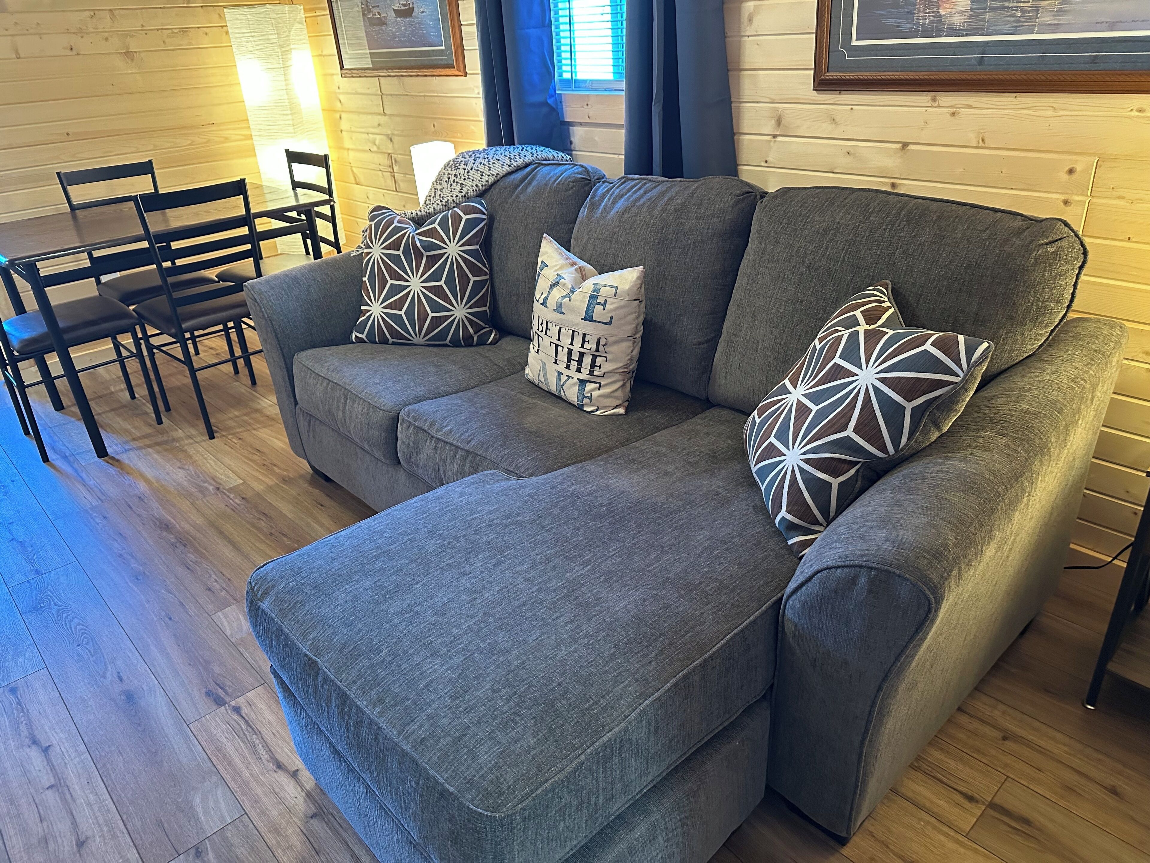 Un canapé d’angle gris avec des coussins à motifs, une table de salle à manger en bois avec des chaises et un tableau encadré au mur.