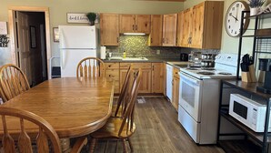 A kitchen with wooden cabinets, a dining table, chairs, a refrigerator, a stove, a microwave, and a clock on the wall. - Perfect 3-bedroom cabin with AC in pleasant Moravia (Moravia)