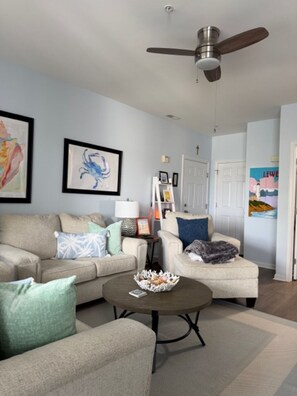 A living room with a ceiling fan, two sofas, a coffee table, and wall art. - Cape Escape! Renovated 2 bedroom/2 bathroom condo! (Lewes)