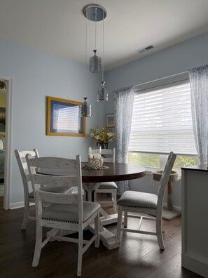 A dining area with a round wooden table, white upholstered chairs, a hanging light fixture, and a window with blinds. - Cape Escape! Renovated 2 bedroom/2 bathroom condo! (Lewes)