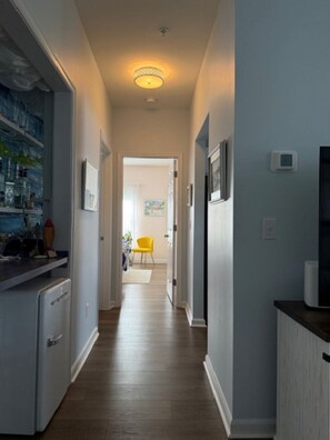 A narrow hallway with a wooden floor, a ceiling light, and a door leading to a room with a yellow chair. - Cape Escape! Renovated 2 bedroom/2 bathroom condo! (Lewes)