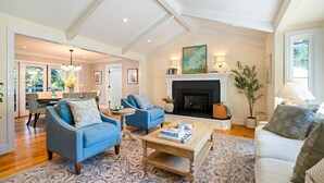 A spacious living room with a fireplace, blue armchairs, a coffee table with books, and a large window. - 4BR Pool Home | Pet-Friendly | Glen Ellen | Near Jack London Park | Sleeps 8 (Glen Ellen)