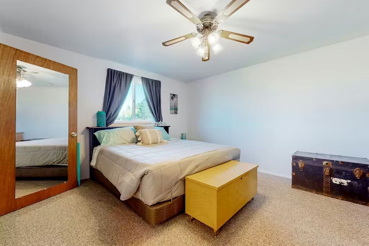 Een slaapkamer met een bed, een plafondventilator, een raam met gordijnen, een nachtkastje en een dressingkast.