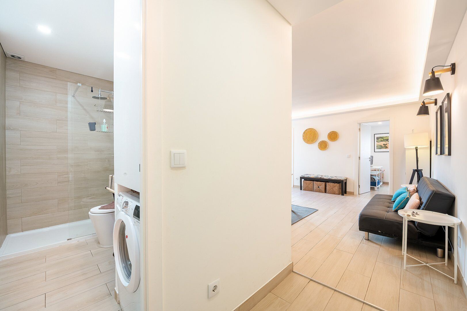 A modern apartment with a laundry area, a living room with a sofa and a small table, and a hallway leading to another room.