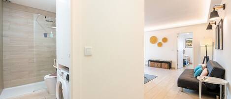 A modern apartment with a laundry area, a living room with a sofa and a small table, and a hallway leading to another room.