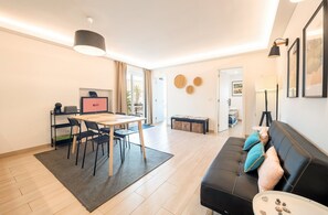 A modern living room with a black leather sofa, a dining table with chairs, a coffee machine, and a TV. - GuestReady - Cosy beachside retreat (Costa da Caparica)