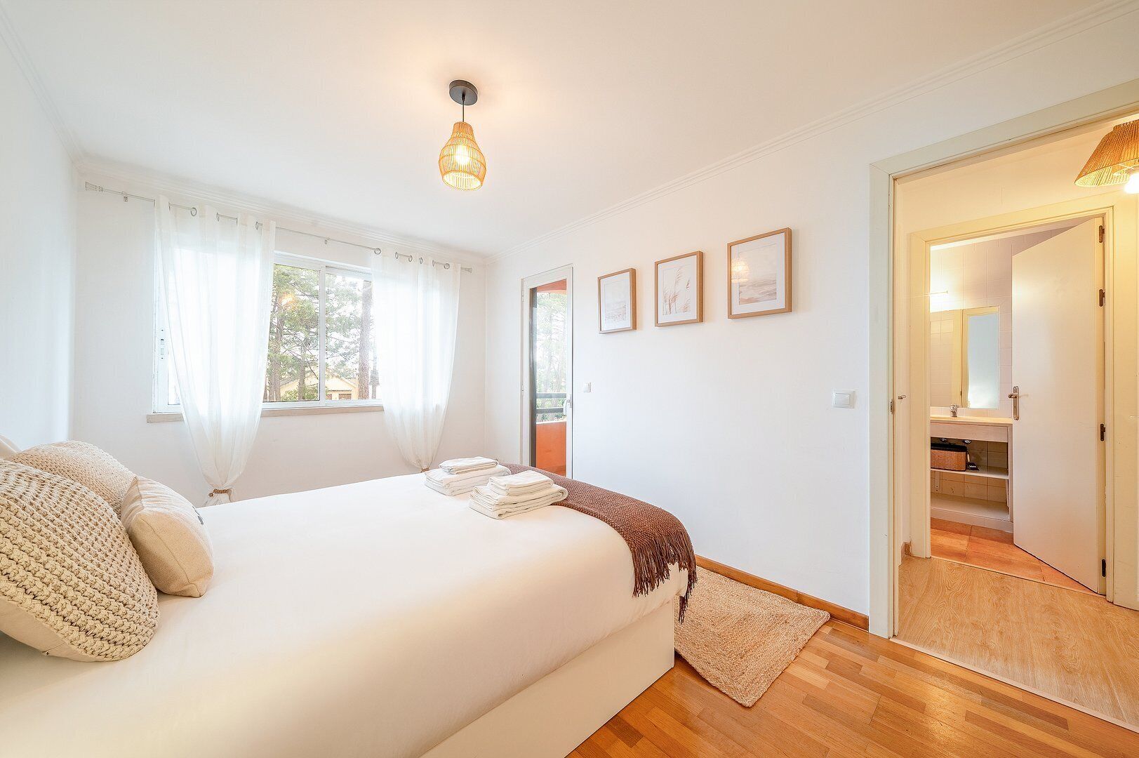 Ein Schlafzimmer mit einem Bett, einem Nachttisch, einem Fenster mit Vorhängen, einer Tür zu einem Badezimmer und gerahmten Bildern an der Wand.