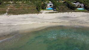 An aerial view of a beach with clear turquoise waters, a sandy shore, and a resort with thatched-roof structures and a swimming pool. - Casa Del Carmen (Santa María Colotepec)