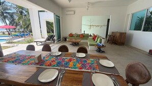 A spacious living area with a dining table set for six, a sofa, and a large window overlooking a pool area. - Casa Del Carmen (Santa María Colotepec)