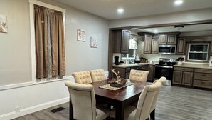 A dining area with a wooden table and chairs, a kitchen in the background, and a window with curtains.