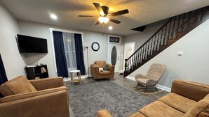 A living room with a sofa set, a rocking chair, a TV, a ceiling fan, and a staircase.
