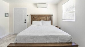 Ein Schlafzimmer mit einem Holzkopfende, einer weißen Bettdecke, einem Nachttisch, einem Fenster mit Jalousien und einer Tür.