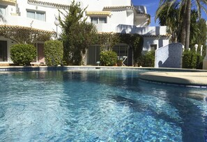 Una piscina con agua azul cristalina, rodeada por un edificio blanco con balcones y palmeras.