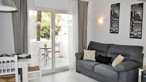 A living room with a grey sofa, a small table, and a chair. There is a sliding glass door leading to an outdoor area with a palm tree and a building in the background. - 06 Ap2116 in Denia With 2 Bedrooms and 1 Bathrooms (Denia)
