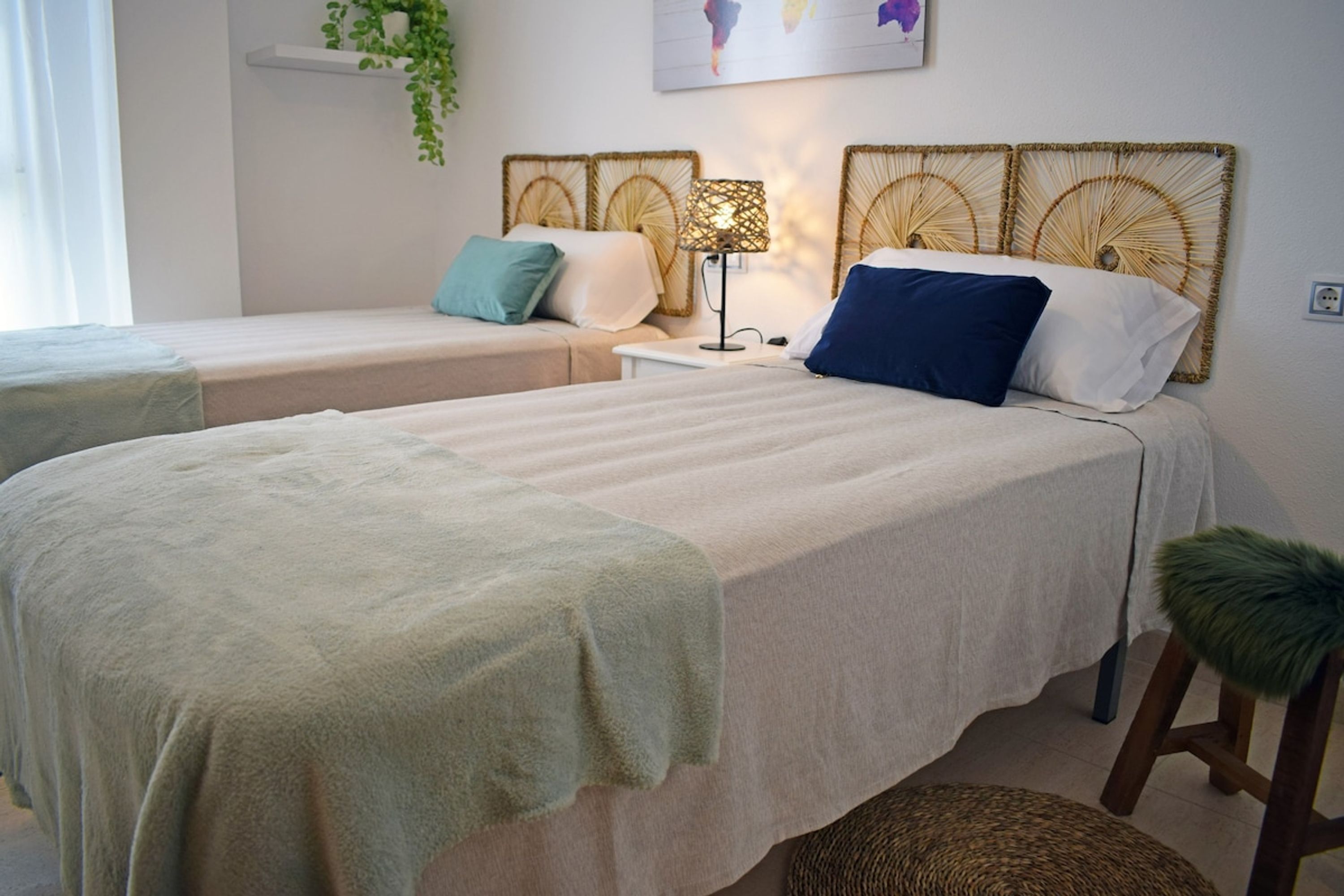A hotel room with two beds, a bedside table with a lamp, a small stool, and a wall-mounted shelf with a plant.