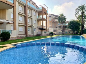 Una piscina con bordo piastrellato, circondata da un edificio con numerosi balconi e un'area giardino con palme.